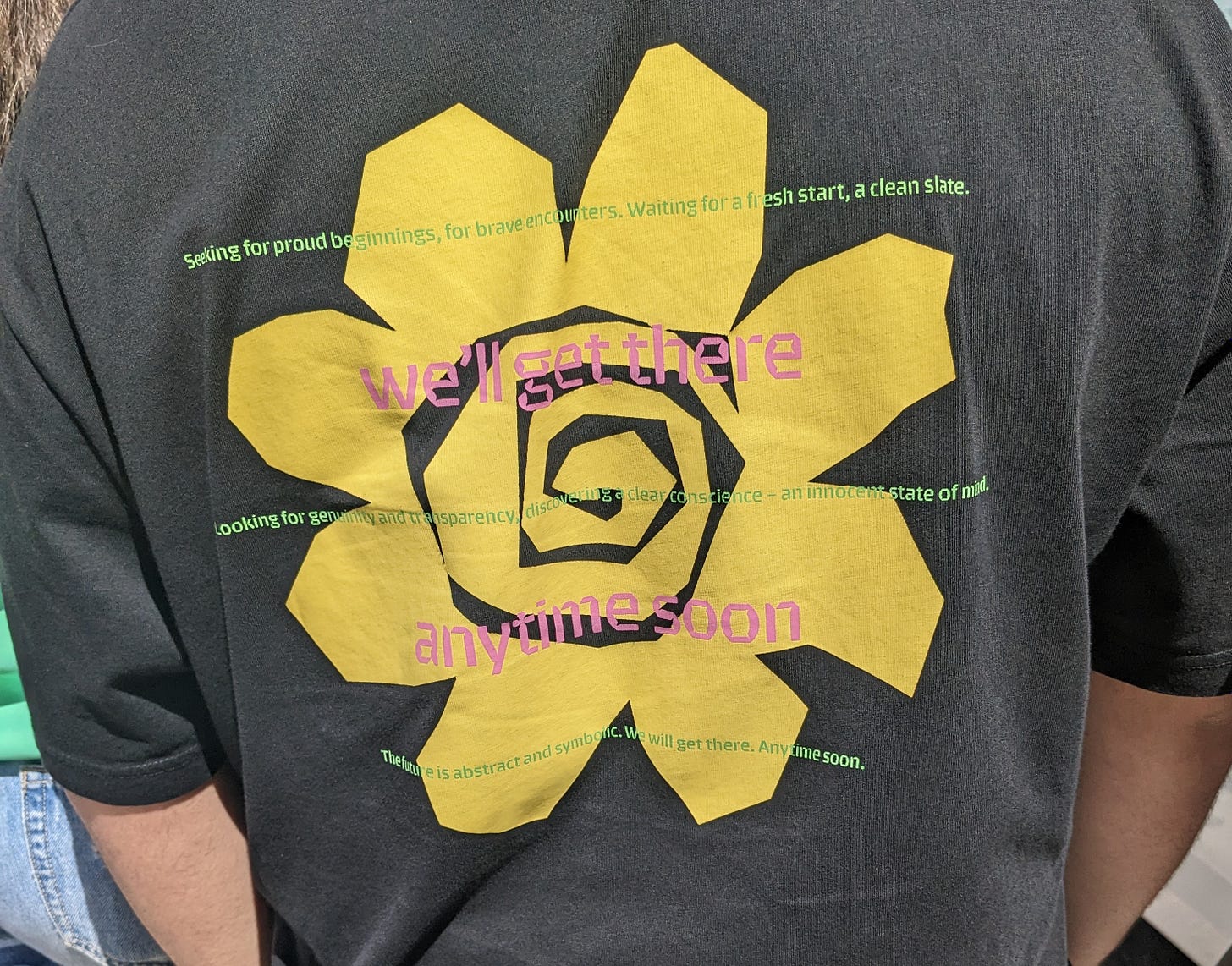 A shirt with a yellow flower and the words Seeking for proud beginnings, for brave encounters.  Waiting for a fresh start, a clean slate.  Looking for genuinity and transparency, discovering a clear conscience – an innocent state of mind.  The future is abstract and symbolic. We will get there. Anytime soon.