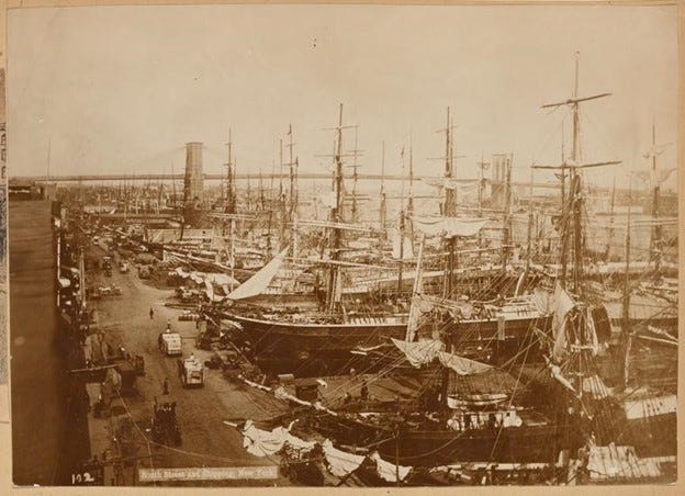 Ships are packed along docks going out into the distance, with the Brooklyn Bridge in the background. A few horse carts, carriages, and pedestrians travel along the street to the left.