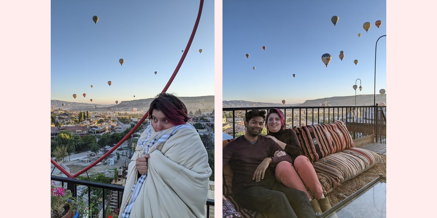 Dawn in Cappadocia, pictured from the rooftop of our bedrock hotel. Me wearing four blankets with balloons rising in the background, and Aseef and I huddled on printed pillows near the railing, with many balloons seen in the air