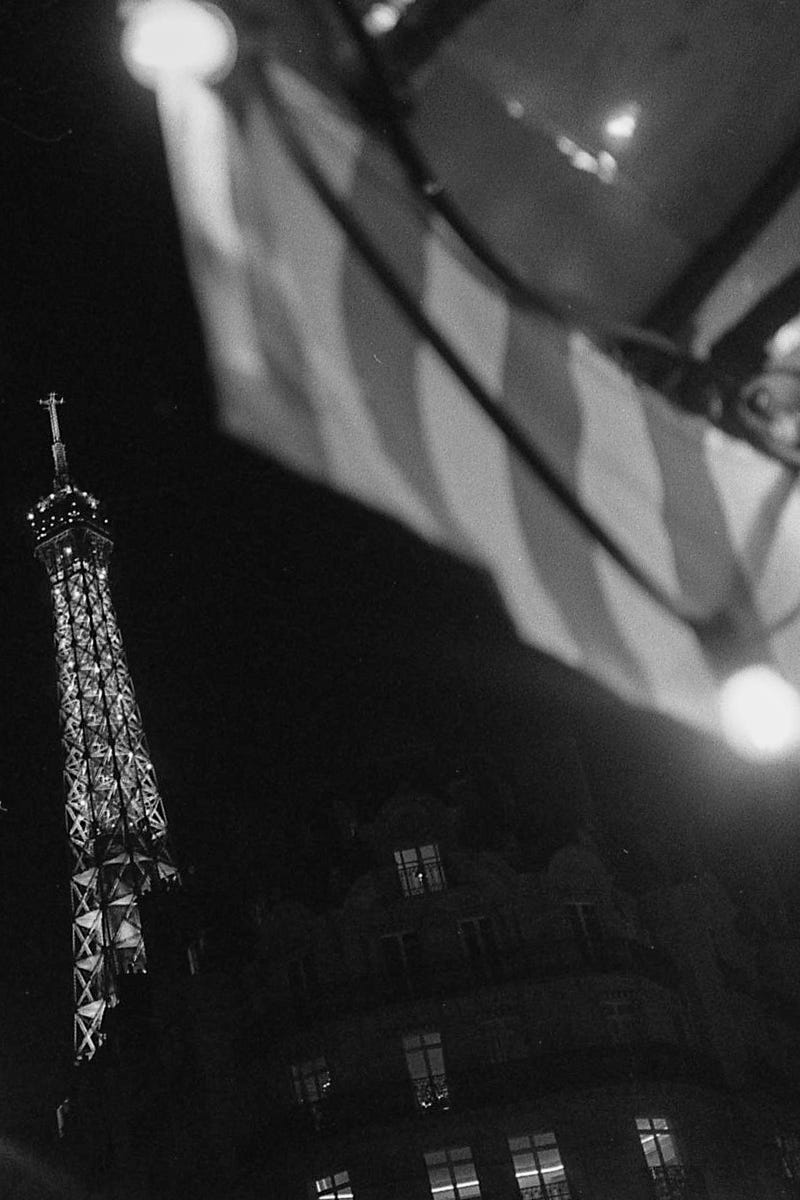 A black and white photograph of the Eiffel Tower lit up at night taken from under a striped restaurant awning with bare lights