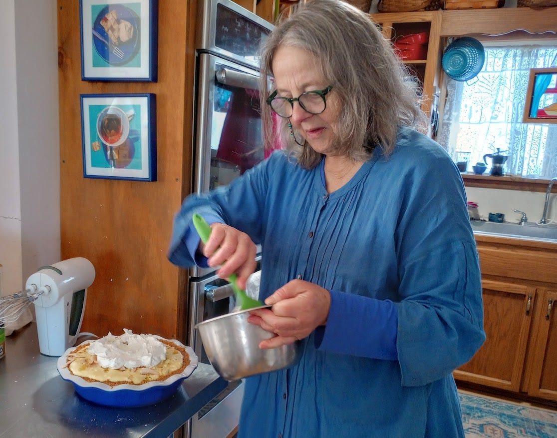 Woman scraping cream from bowl with spatula, to place on pie sitting on table, in kitchen