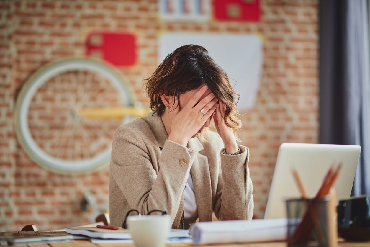 A young woman puts her face in her hands while sitting at her desk in a modern office.