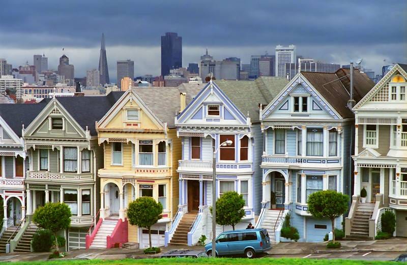 Six colorful victorian homes in the foreground. On the street level, each has a garage. The San Francisco city skyline is in the background.