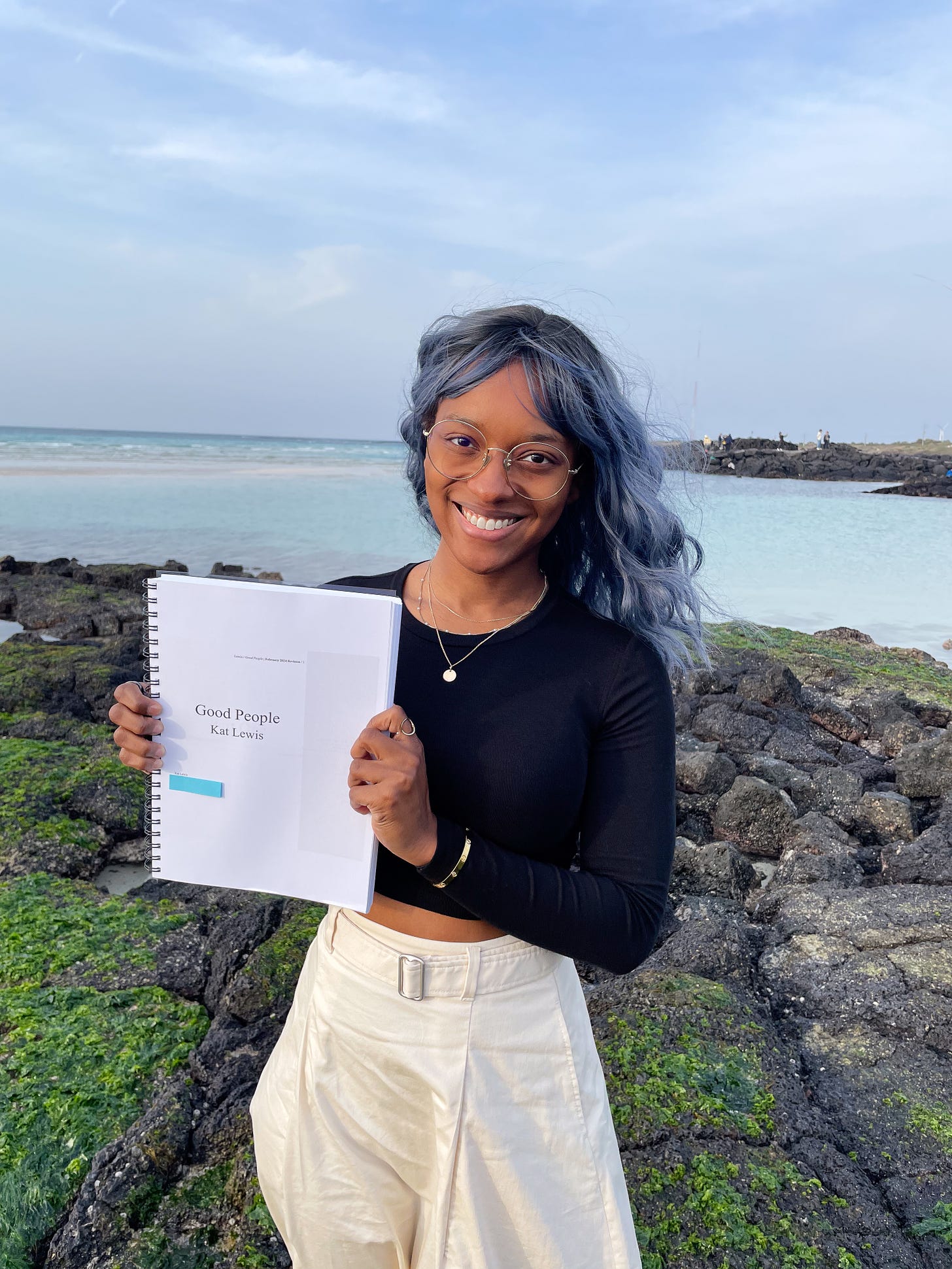 A photo of Kat Lewis, a Black woman with blue hair and large round glasses. She stands on a volcanic rock beach on Jeju Island in South Korea. She holds a printed manuscript. The title page says, “Good People by Kat Lewis.”