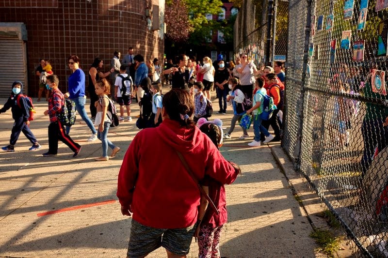 A woman with a red coat has her arm around a girl outside a schoolyard.