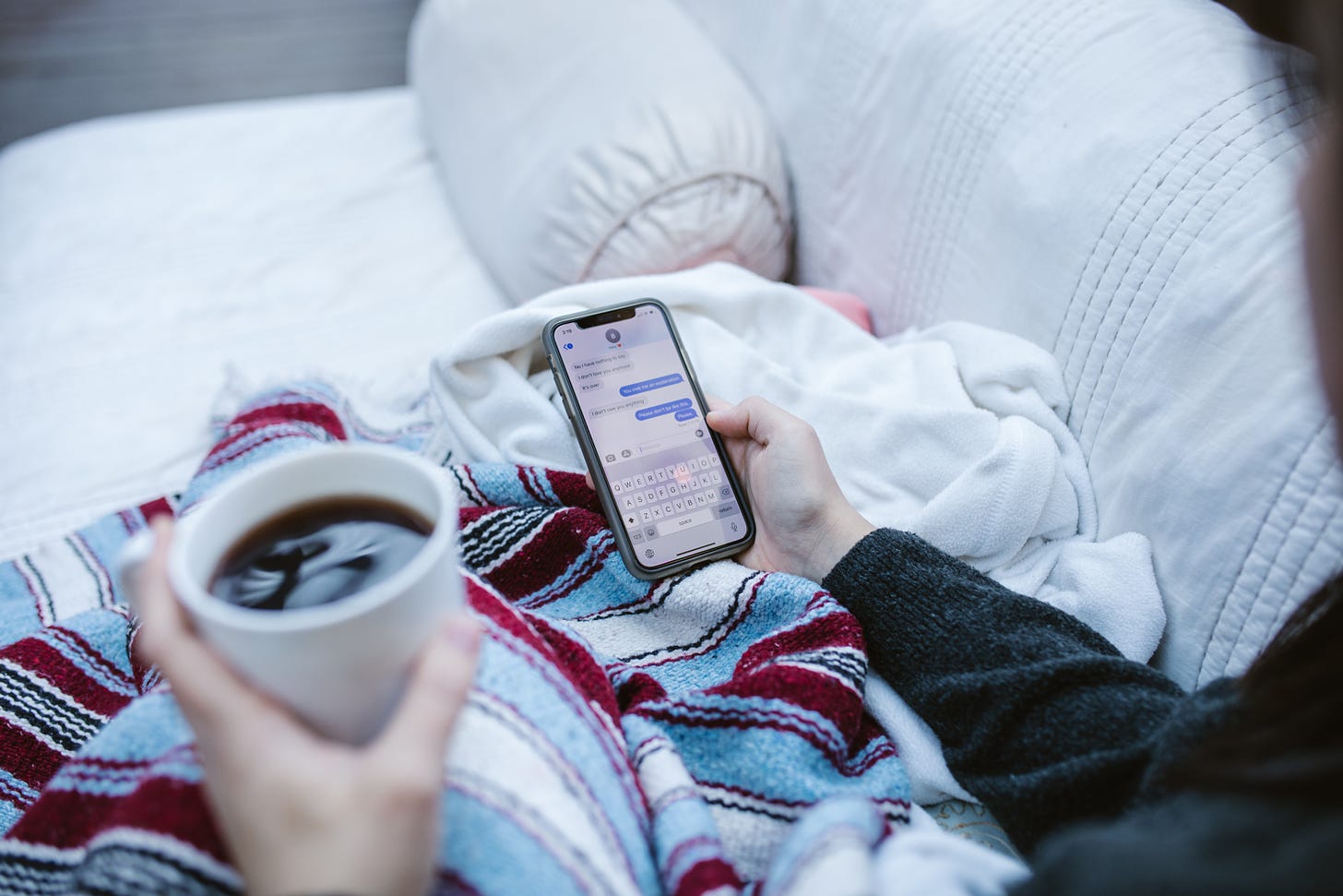 a person sits on a sofa in a blanket, reading text messages on an iphone