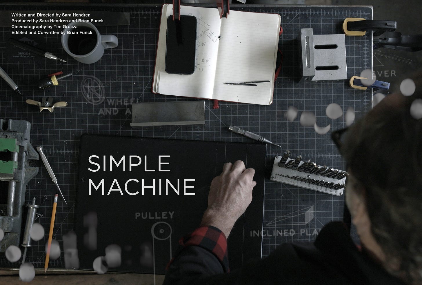 An overhead shot of a man at a workbench with tools, coffee mug, phone and notebook strewn about and the title Simple Machine and credits in white.