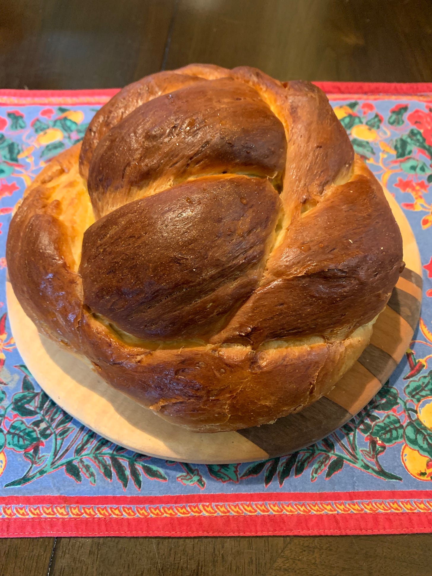 A challah, beautifully browned, set on a round cutting board on top of a blue placemat with a floral motif. 