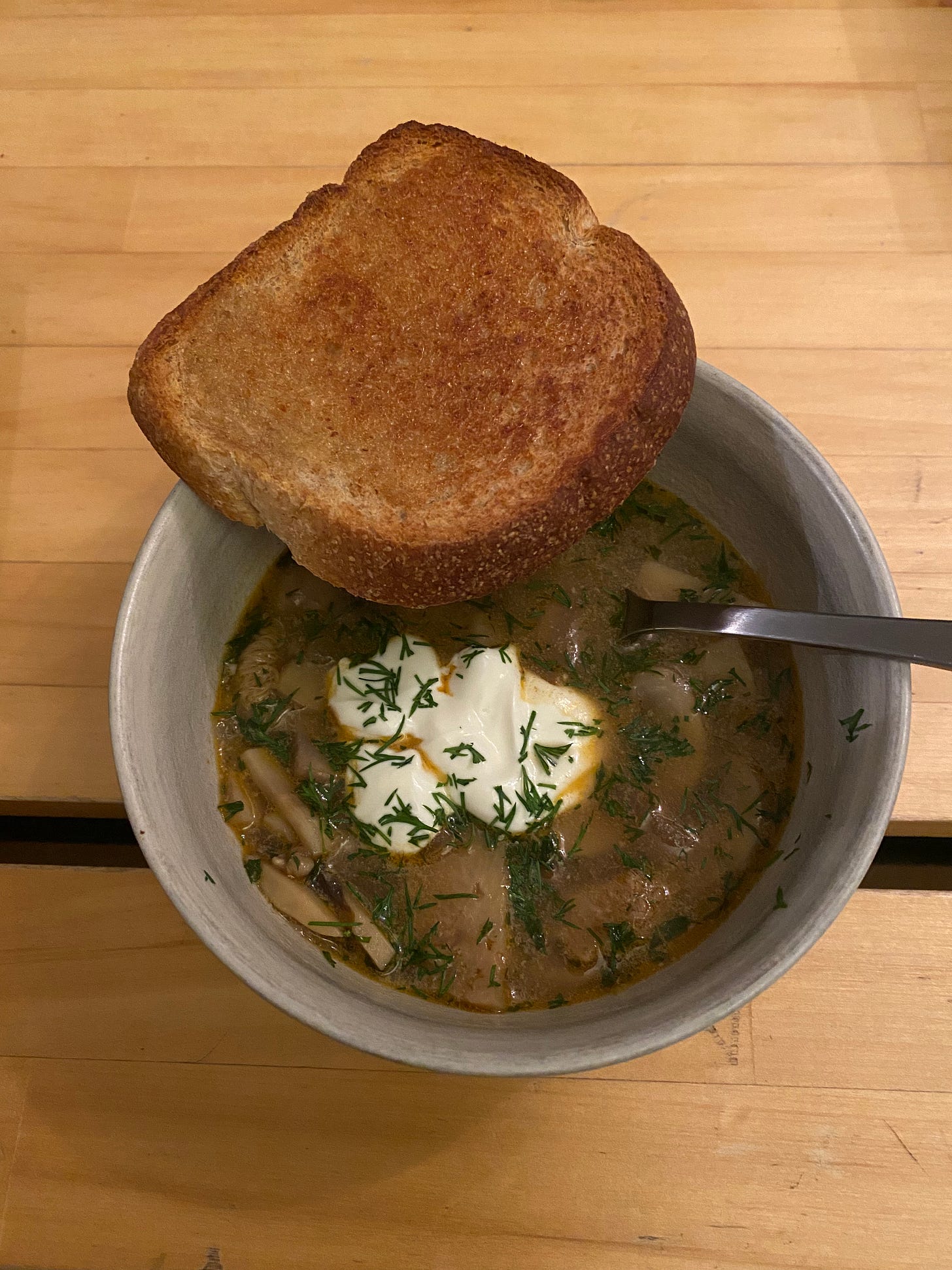 A white bowl of an orange-brown mushroom soup flecked with dill and topped with sour cream. A slice of buttered wheat toast rests on the edge.