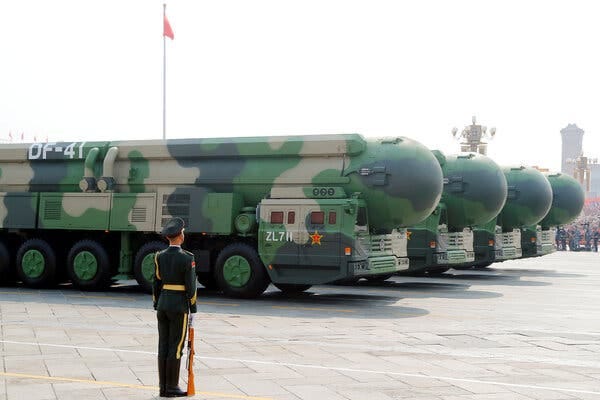 A Chinese soldier standing in front of 4 large ballistic missiles painted in camouflage green during a military parade.