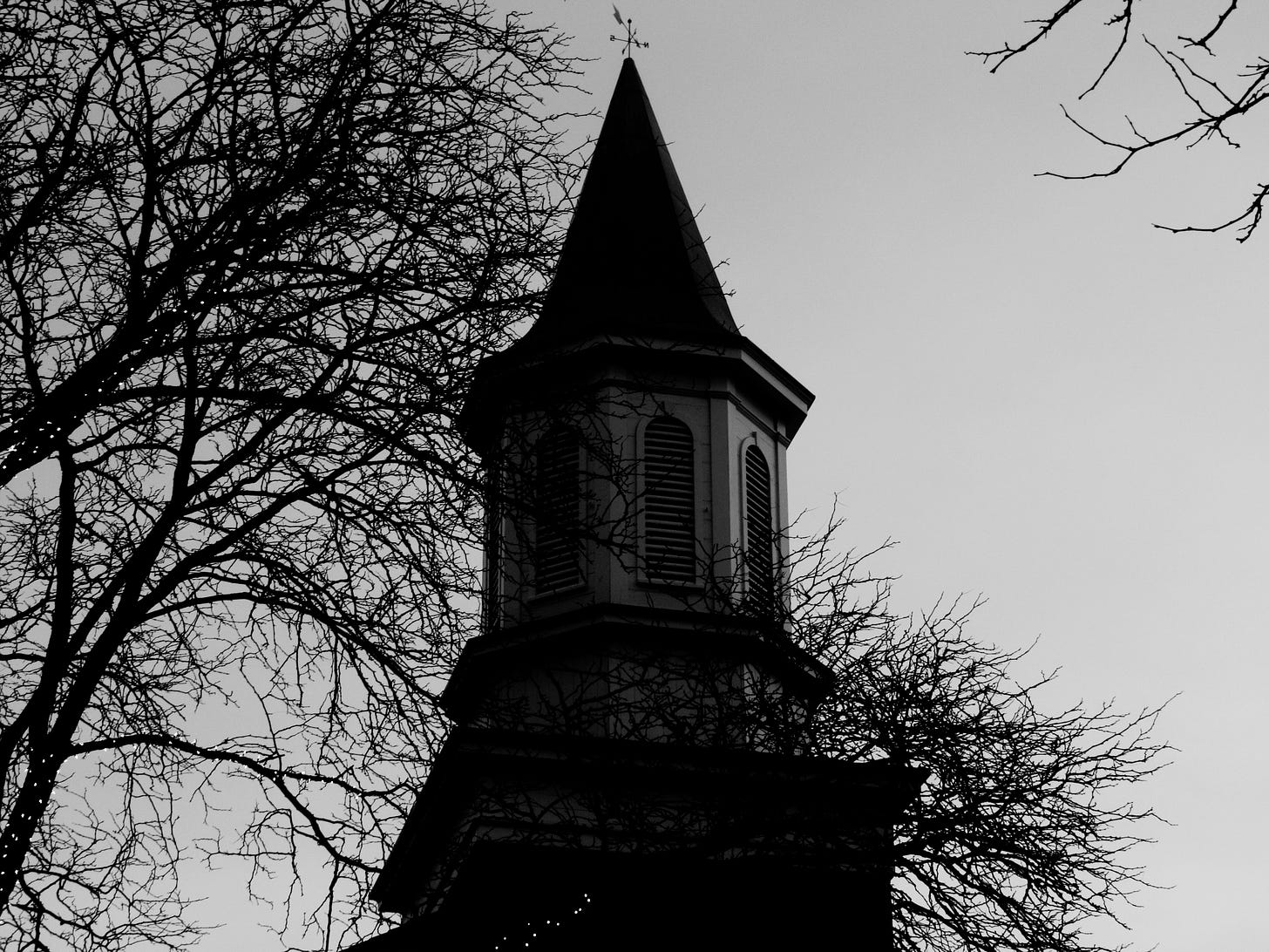 A black and white photo of a church's steeple with a weather vane on the pea and a leafless tree next to it