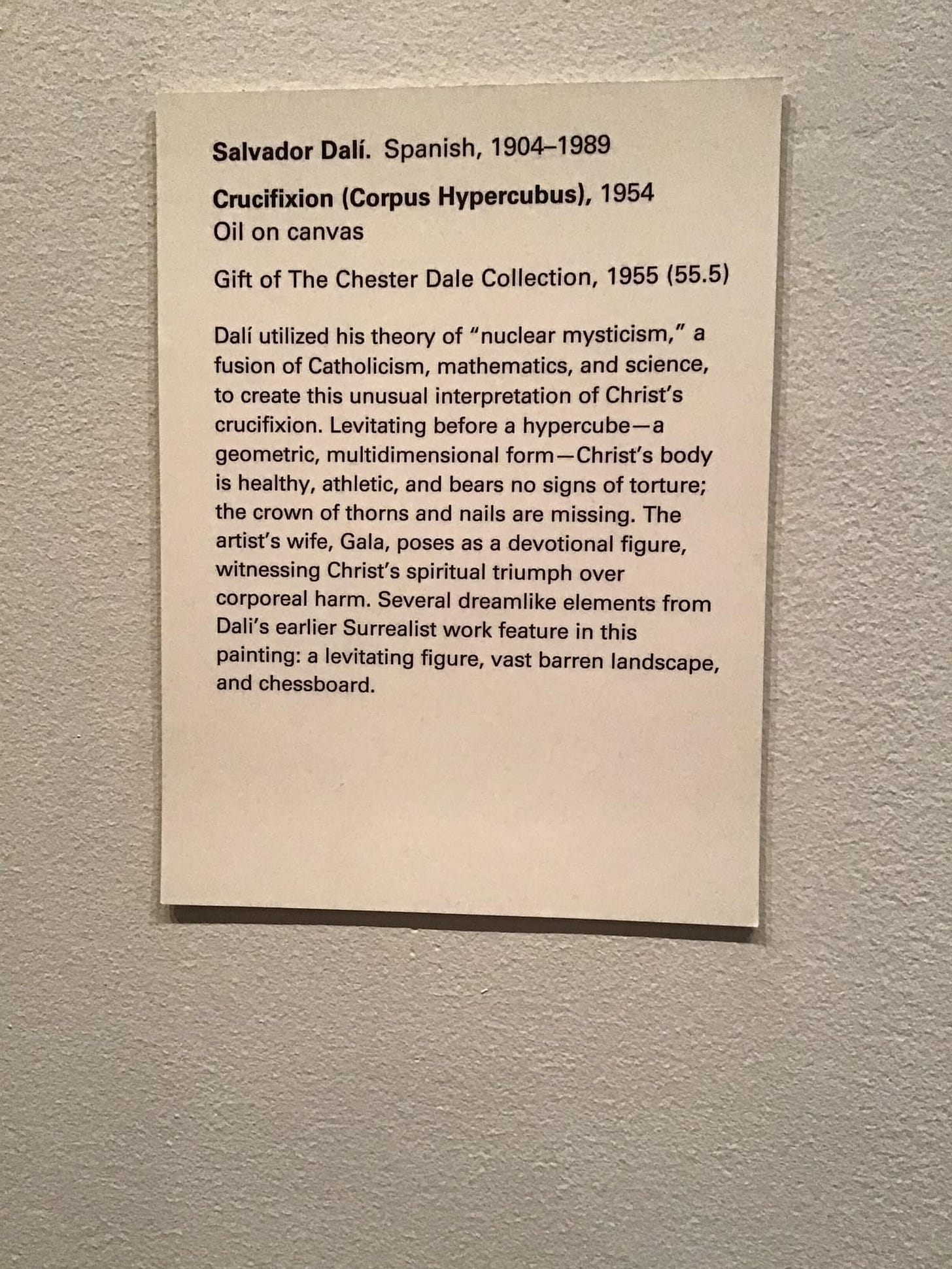 May be an image of text that says 'Salvador Dalí. Spanish, 1904-1989 Crucifixion (Corpus Hypercubus), 1954 Oil on canvas Gift of The Chester Dale Collection, 1955 (55.5) Dali utilized his theory of "nuclear mysticism, a fusion Catholicism, mathematics, and science, create this unusual interpretation of Christ's crucifixion. Levitating before hypercube-a geometric, multidimensional form- -Christ's body is healthy, athletic, and bears no signs of torture; the crown of thorns and nails are missing. The artist's wife, Gala, poses devotional figure, witnessing Christ's spiritual triumph over corporeal harm. Several dreamlike elements from Dali's earlier Surrealist work feature in this painting: levitating figure, vast barren landscape, and chessboard.'