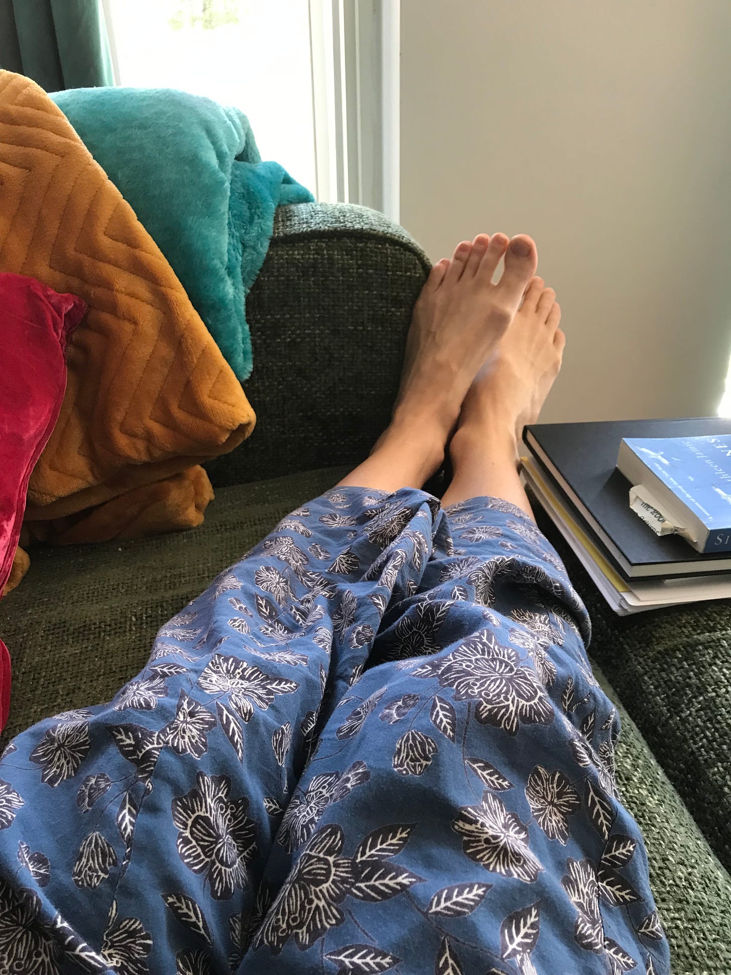 view of my legs in blue pyjamas and bare feet on a sofa with turquoise and mustard velvet blankets folded up and books to the side.