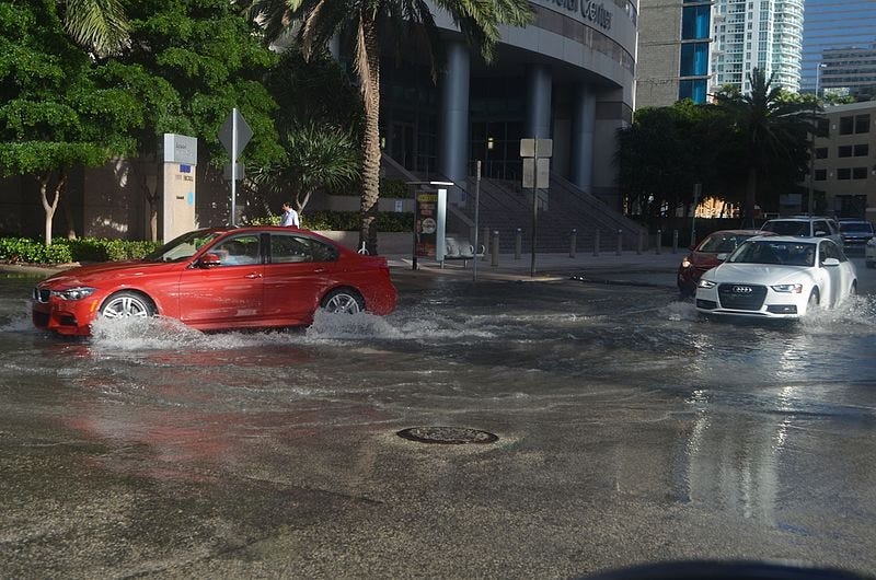 During a 'king tide,' a red BMW and white Audi drive through several inches of seawater flooding streets on a sunny day in downtown Miami