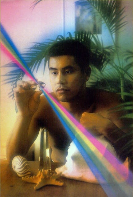 An Asian man is sitting down, and there are large shells on the table near him. He is holding a crystal, which is shining a rainbow light diagonally across the image. There is a green plant behind him, and a framed artwork on the white wall behind him. An Asian man is sitting down, and there are large shells on the table near him. He is holding a crystal, which is shining a rainbow light diagonally across the image. There is a green plant behind him, and a framed artwork on the white wall behind him.