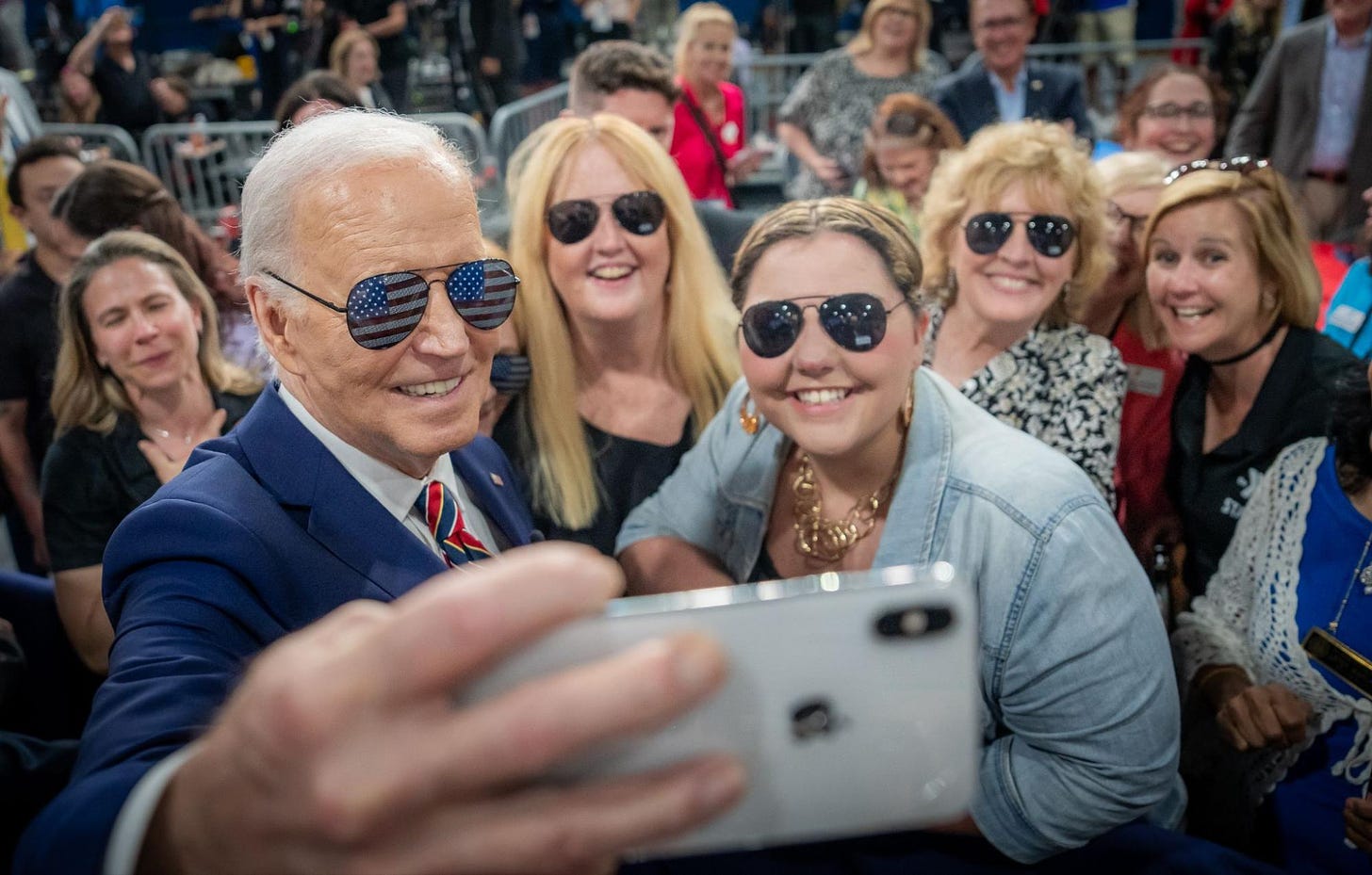 President Joe Biden takes a selfie with a group of event attendees. President Biden has on a pair of aviator sunglasses with the American flag in the lenses. The women in the selfie have on black aviator sunglasses.