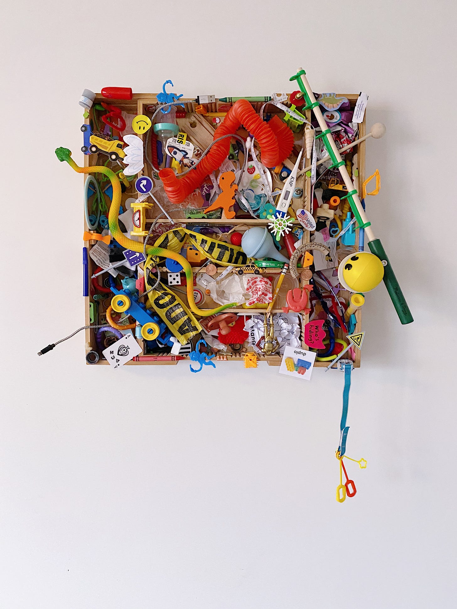 Broken toys and detritus are glued to a bamboo silverware tray. Items include: plastic snake, pop tube, caution tape, barrel of monkeys, smiley face sticker, dum dums wrapper, I voted sticker, children's fishing pole, crayons, broken cars, and more.