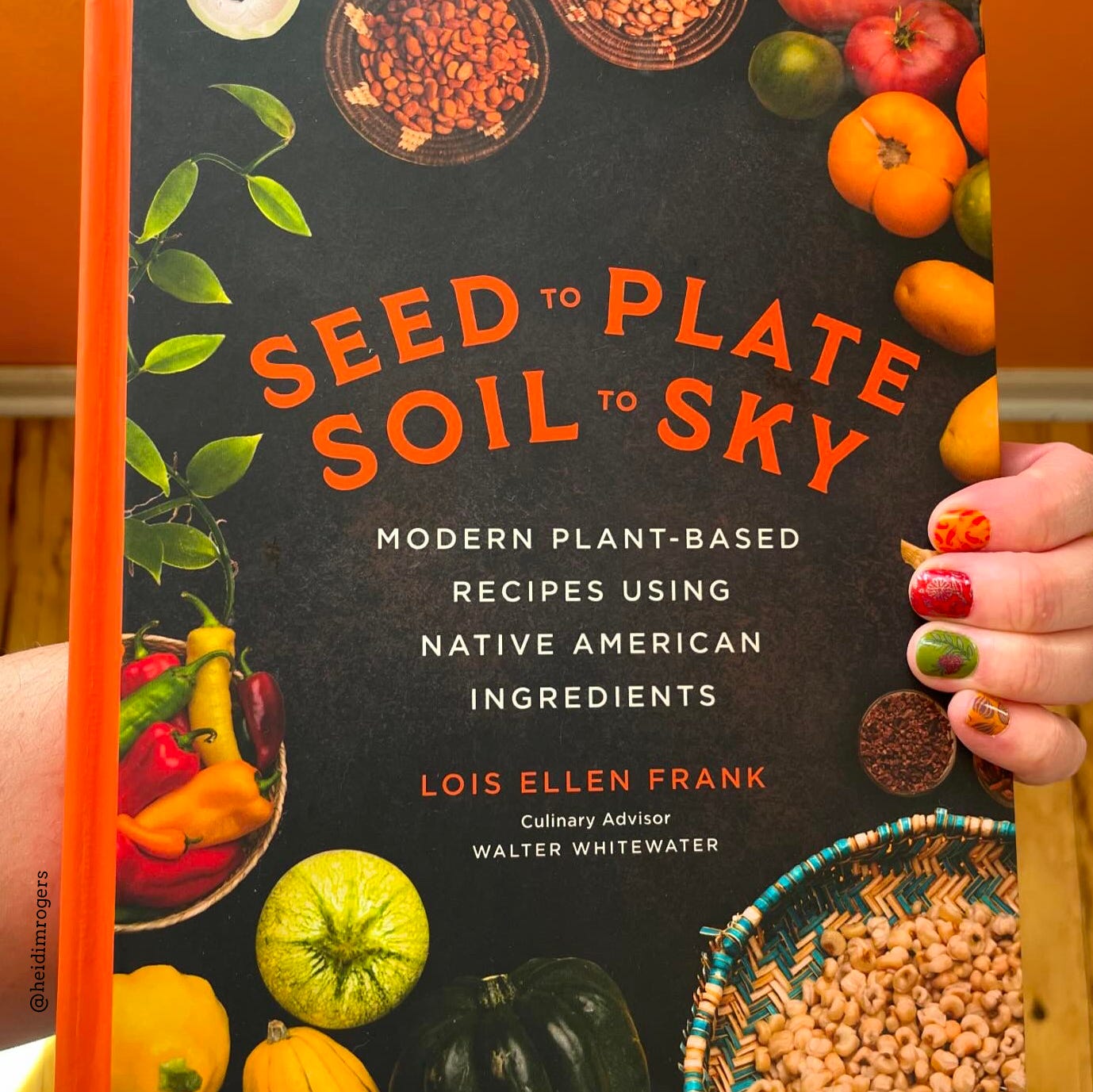 White woman's hand is holding the cookbook, Seed to Plate Soil to Sky, over a wood floor and orange wall background.