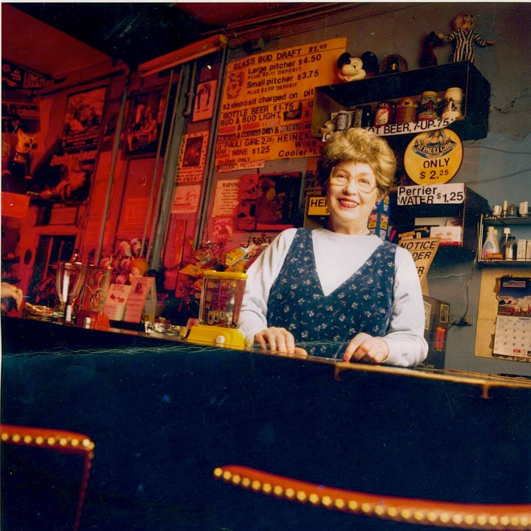 Bartender in the 90s with brown hair and a pretty smile. She is white and looks like someone you might see at church on Sunday. Brown wig. Dive Bar.