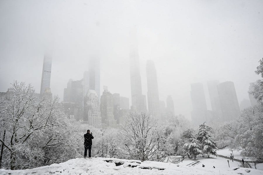 A person takes a picture in a snow-covered park with skyscrapers standing among low clouds in the background.