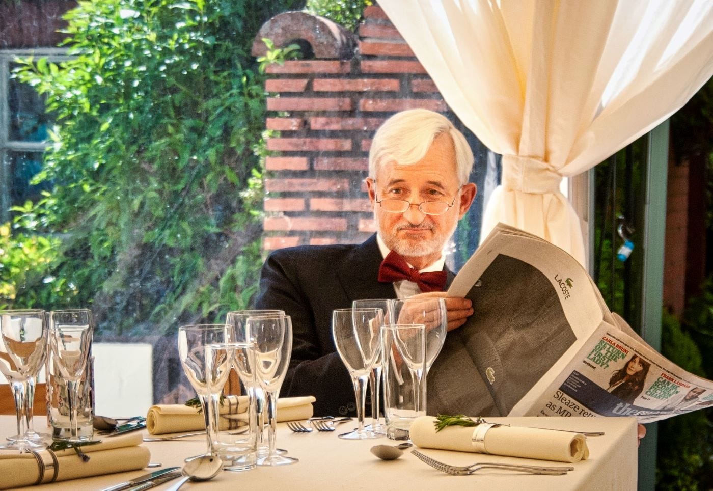 A man with white hair and a beard, wearing a suit and bowtie, is reading the newspaper at a table laid in a garden. He is looking up from reading the newspaper, with his reading glasses perched on his nose.