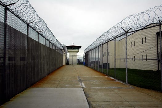 Photo of cement walkway with tall fences and barbed wire on either side--prison
