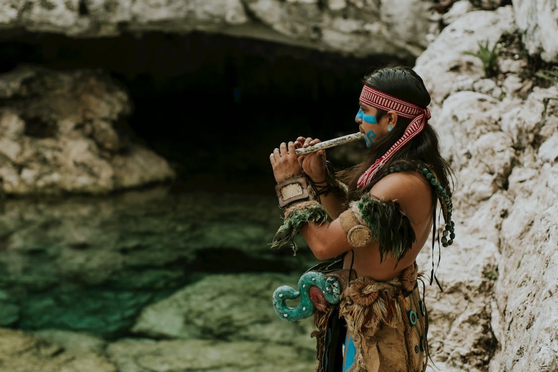 Free Man in Traditional Maya Clothes and Face Paint Standing by Water Playing Instrument Stock Photo