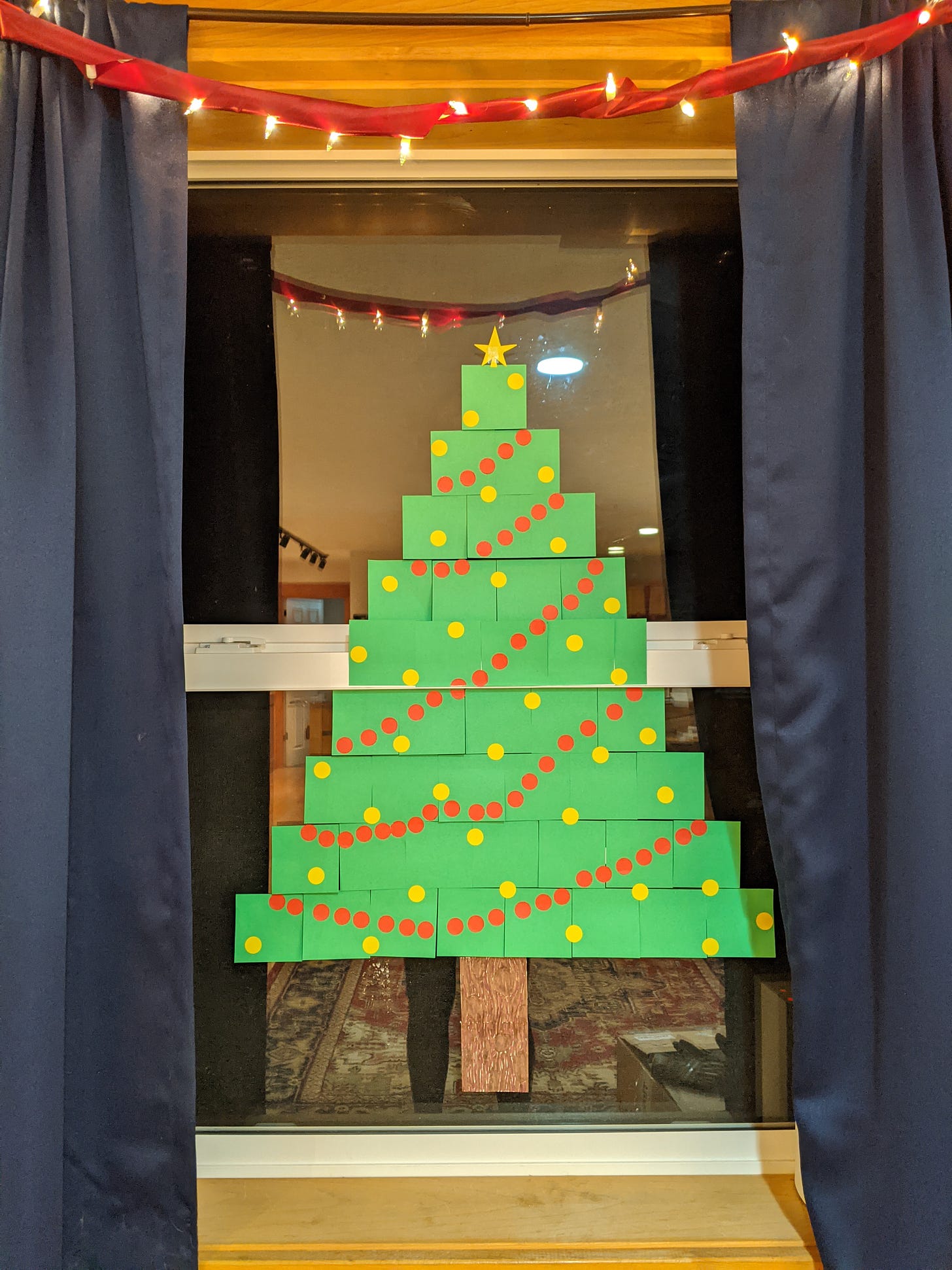 a paper christmas tree made of green post-it notes and yellow and red stickers arranged like a garland and ornaments, all stuck to a window