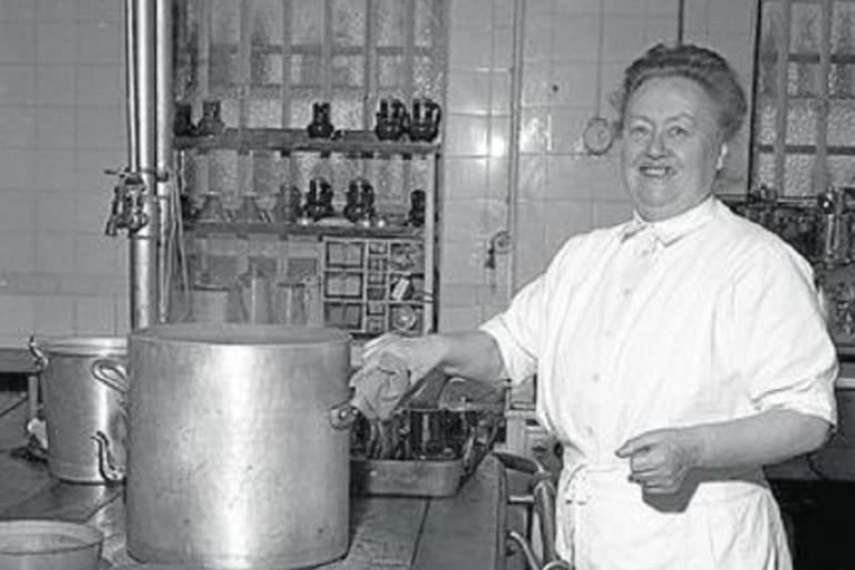 Black and white vintage photo of Eugenie Brazier in what looks like a restaurant kitchen. She's smiling at the camera as she tens to a large stockpot on the stove. Image source: Al Jazeera (https://www.aljazeera.com/features/2018/6/12/eugenie-brazier-why-google-honours-her-today)