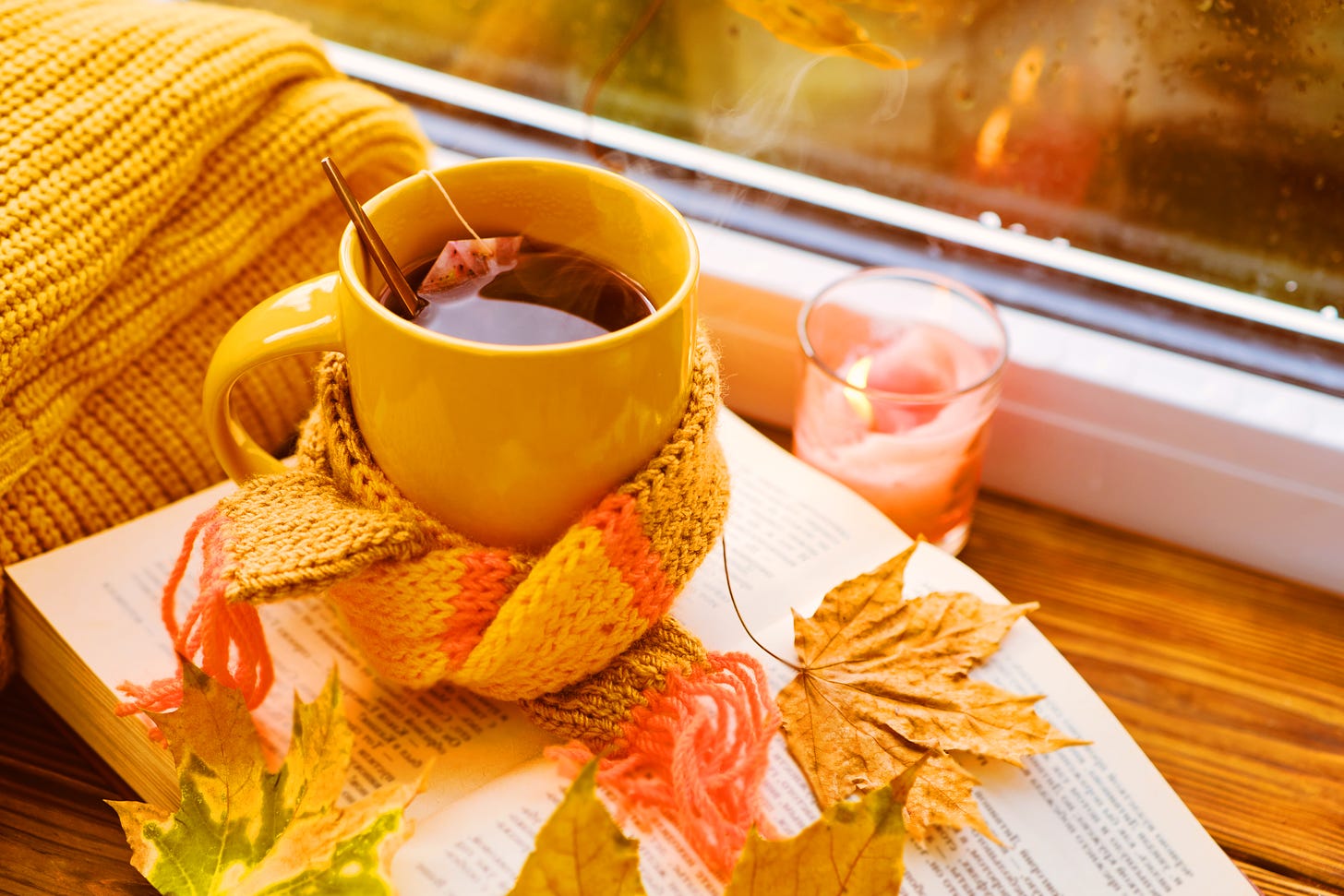 Close-up of a windowsill where there’s a cup of hot tea with a orange and yellow striped scarf, over an open book, beside a little candle. There are dried leaves scattered over the book as well.