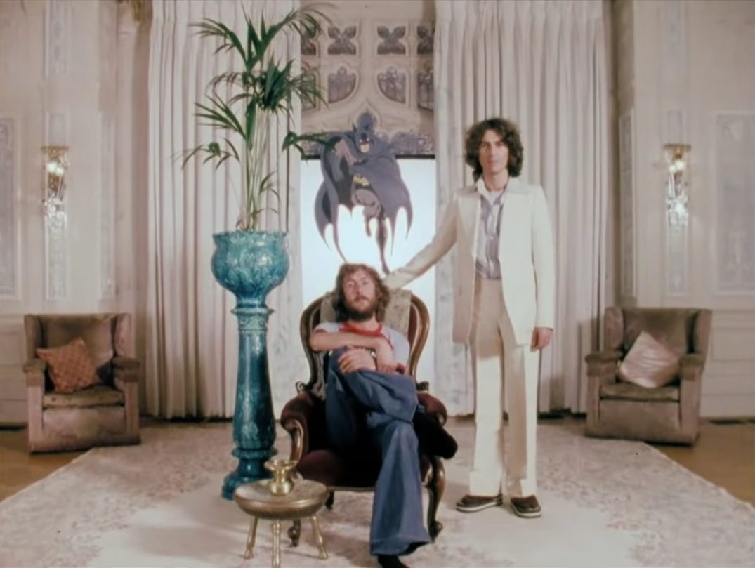 Eric Idle sits in an armchair, wearing a t shirt and jeans, while George Harrison stands beside him, wearing a white suit