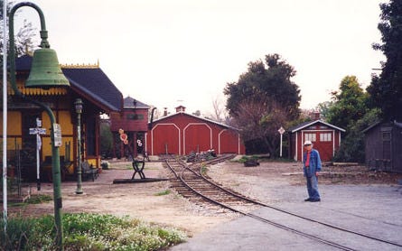 An elderly man standing next to railroad track in the foreground with an old-fashioned railroad depot building on the opposite side. A locomotive shed and water tower are located where the railroad track ends in the background.