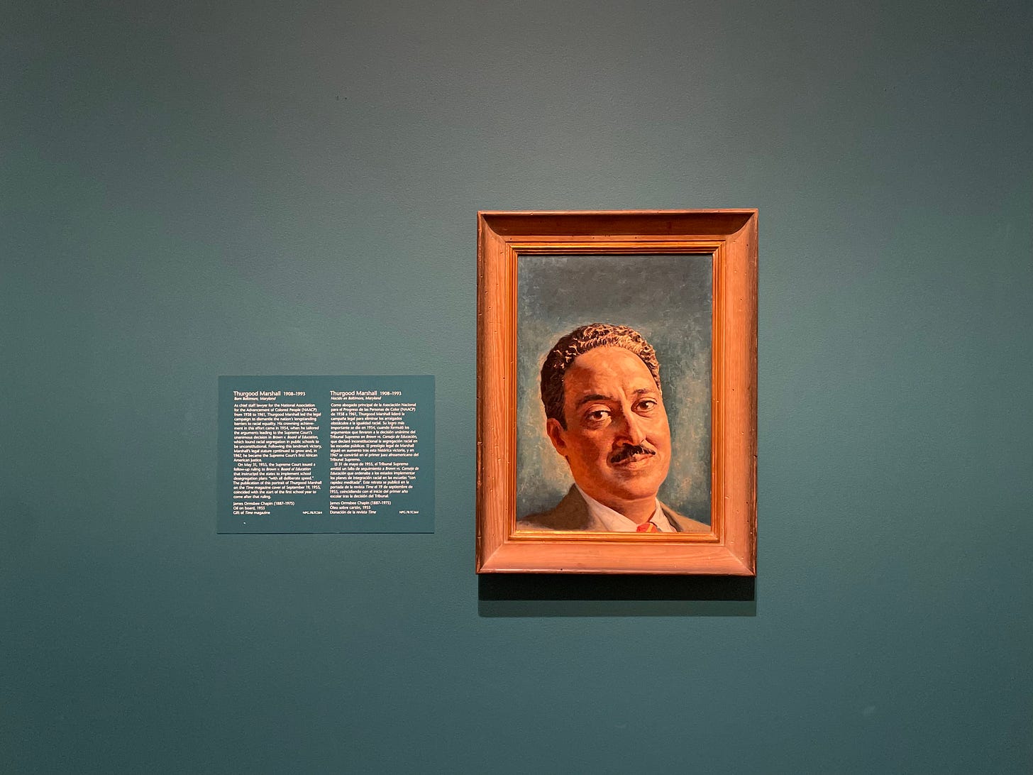 This is a photograph of a portrait painting of Thurgood Marshall. Marshall is depicted as a middle-aged Black man with short dark hair and a moustache. He is dressed in a suit and tie. The painting’s background is teal, as is the wall on which the painting hangs.