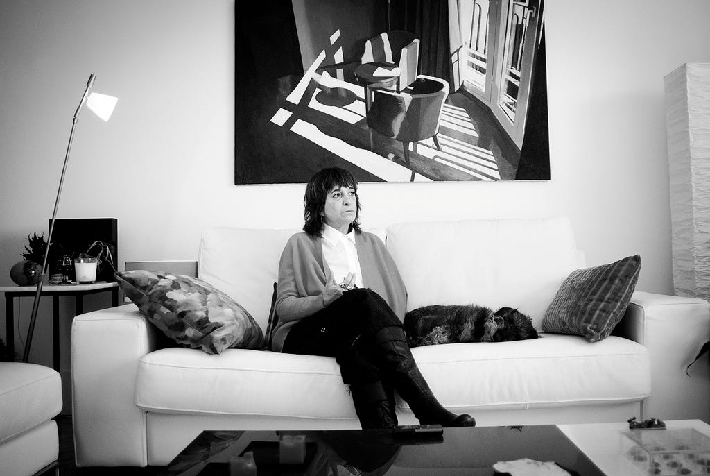 Rosa Montero sentada num sofá, ao lado de um cachorro deitado, numa sala de estar. ela é uma mulher branca, de cabelos escuros, nos ombros.