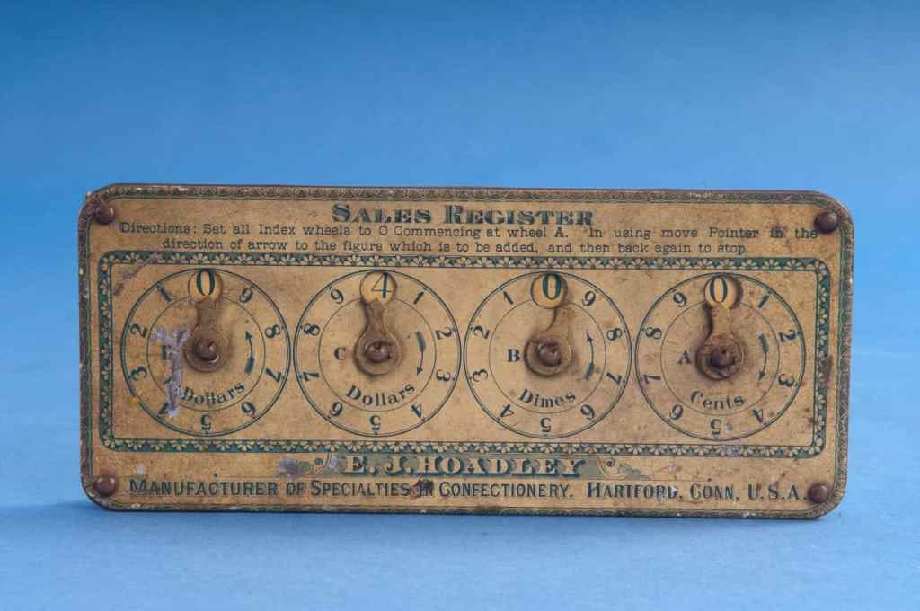 This small device has a tin case painted yellow and green on top and red around the edge. The top of the case has four wheels drawn on it, each of which has the digits from 1 to 9 drawn around the edge. The digits go clockwise for the first and third circles (marked “Cents” and “Dollars” and counterclockwise for the second and fourth circles (marked “Dimes” and “Dollars”). At the top of each circle, at the zero position, is a window that reveals a rotating disc below. The discs are rotated using thumbscrews that protrude from the back of the instrument. An arm on top of each circle points to a digit on the wheel. The discs advance when they are rotated in the direction of increasing digits and remain fixed when the arrows are moved back to zero.