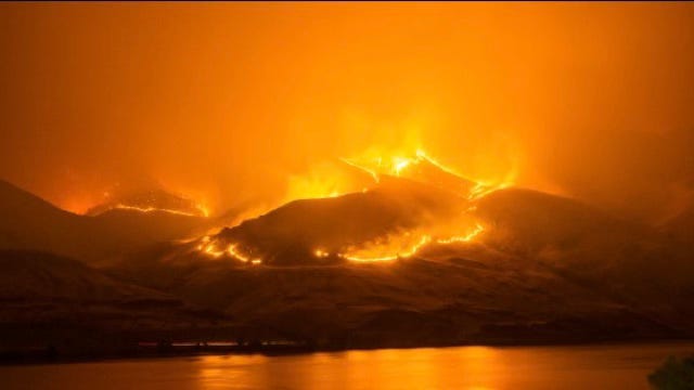 A ring of fire burns around the peaks of a volcano, setting the sky ablaze, as well as its reflection in the water.