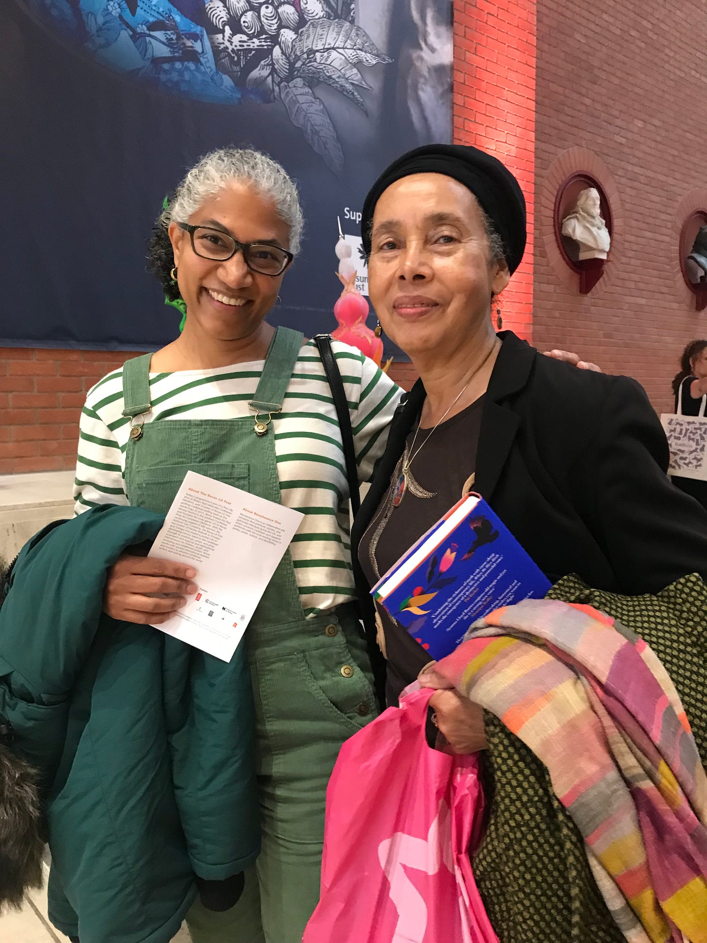 Grace Nichols, on the right, standing with Amanda Coreishy on the left, both holding their coats. Amanda is holding The Bocas UK Tour 2022 schedule and Grace is holding a copy of When We Were Birds