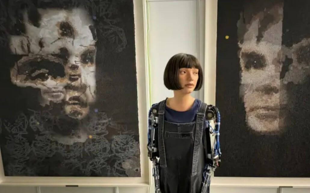 Photo of Ai-Da, a humanoid robot, standing between two portraits of Alan Turing painted by the robot. The portraits display an abstract, pixelated style with Turing's face emerging from dark backgrounds.