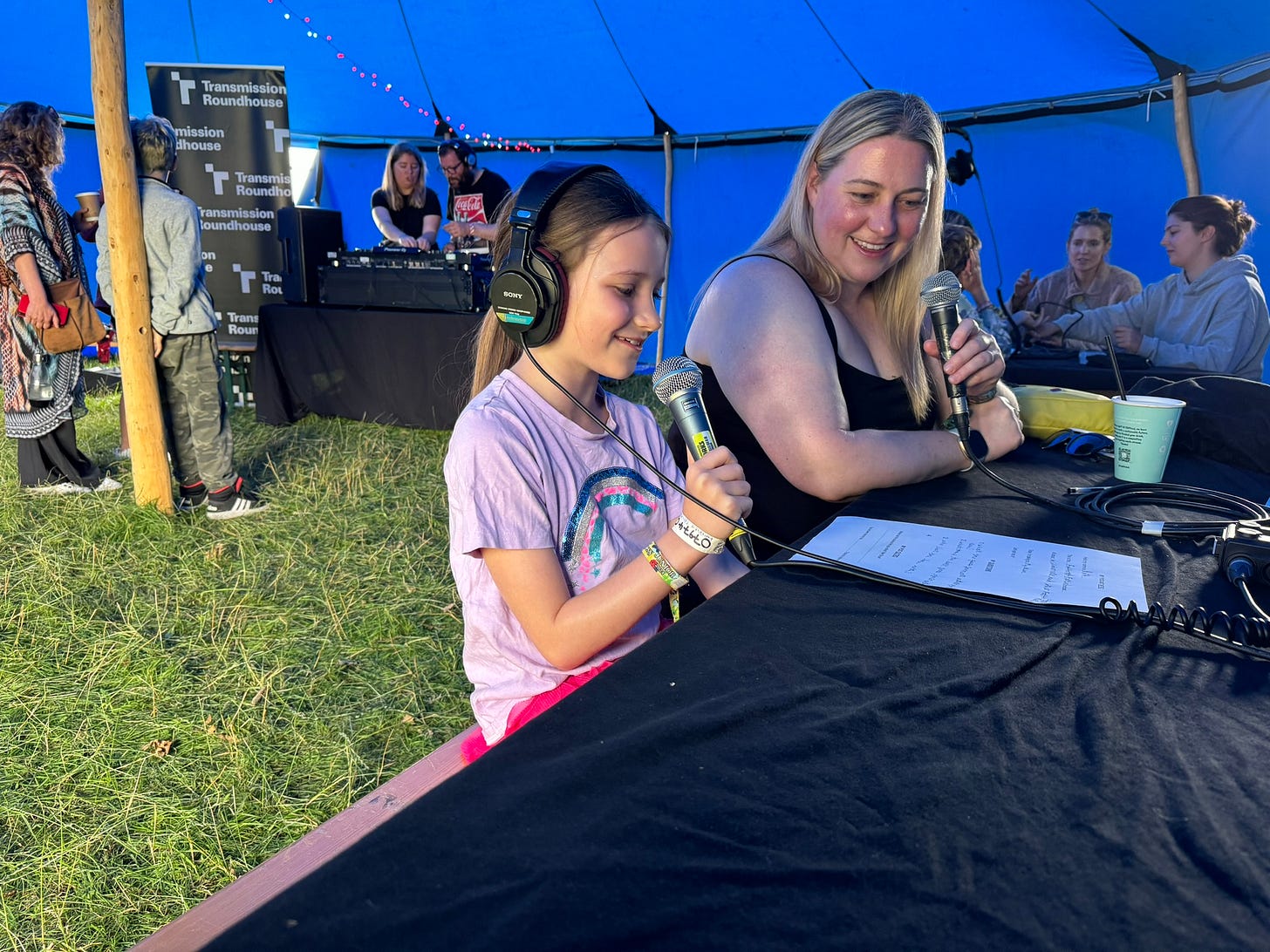 Inside a blue tent are tables around the edges.  Beth is a young girl with brown hair tied back in a pony tail.  She is holding a microphone and has earphones on.  Her mum is blonde and is sat next to her with a microphone in her hand also.  