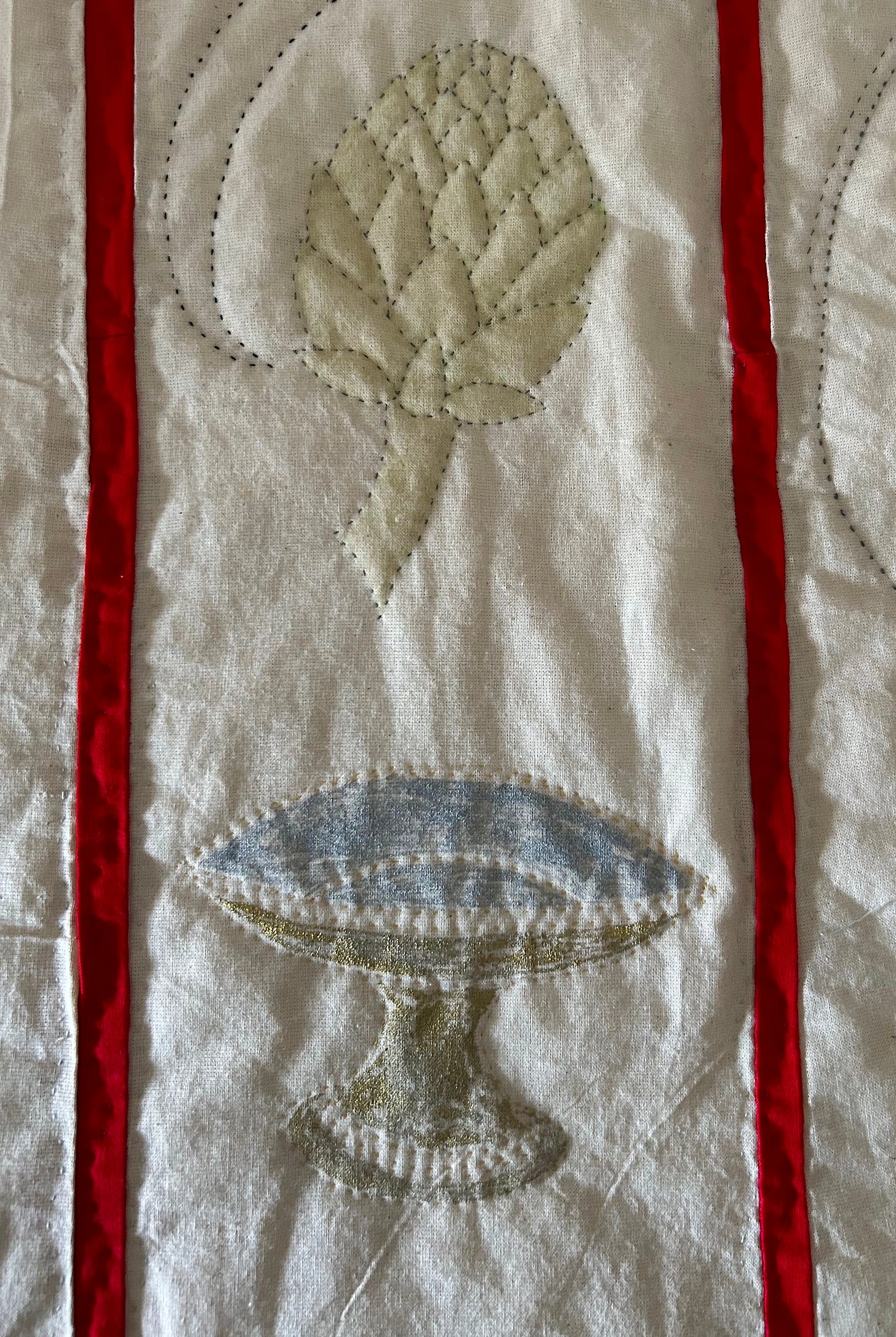 Cream cloth, quilted with a pale green artichoke and a gold and silver dish on a stand. There are two red bands - each on either side of the images depicted.