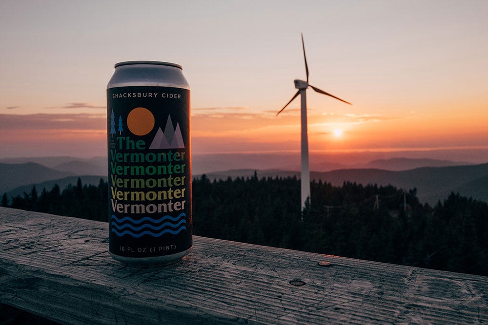 A can of Vermonter cider sits on a wooden railing with a windmill in the background and the sun setting over the mountains.
