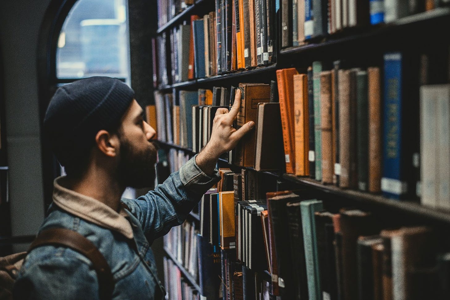 A man wearing a beanie thumbs through titles on a library bookcase