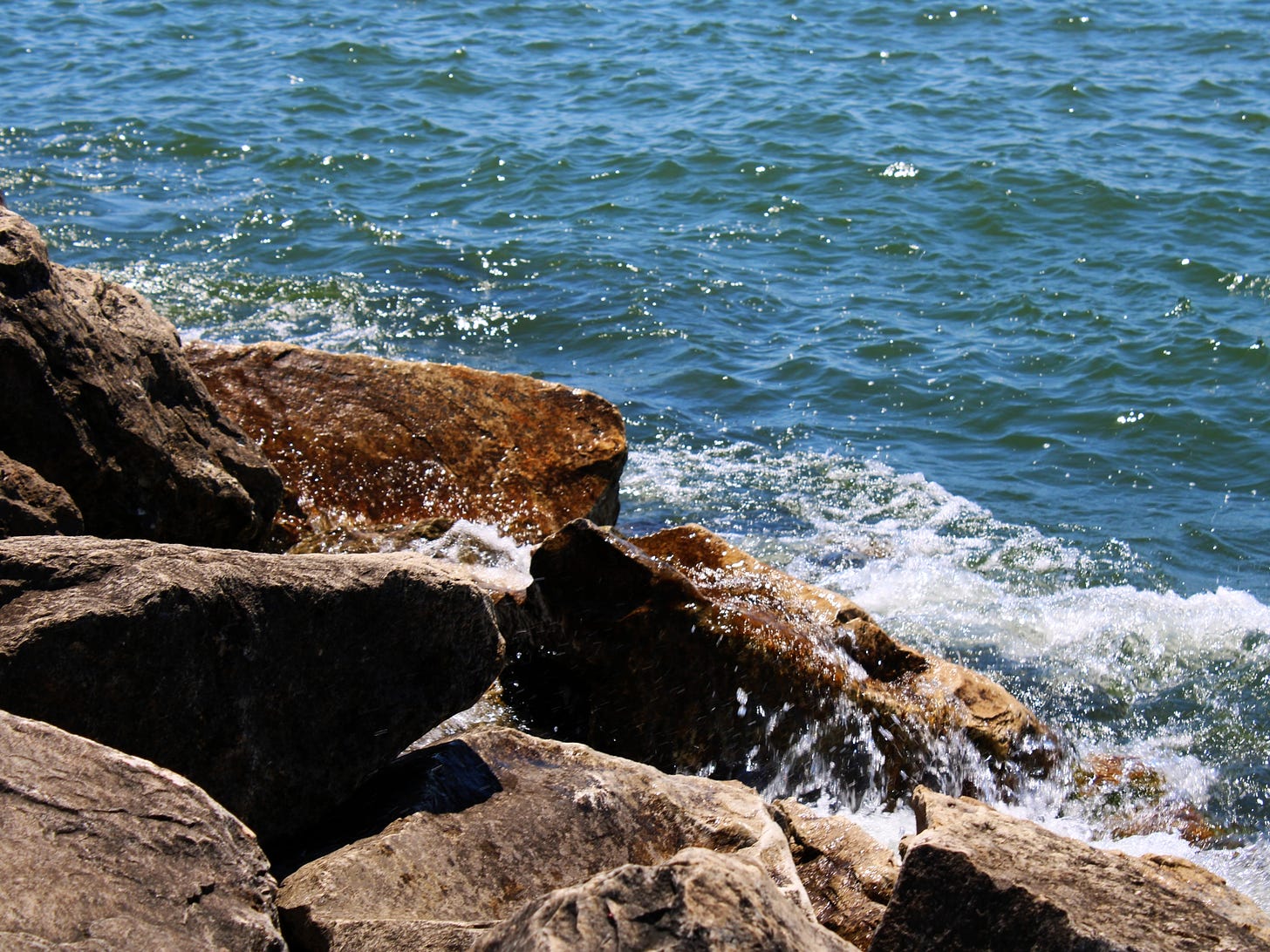 A photograph of brownish gray rocks in the foreground with foamy Lake Michigan water breaking on them in the midground.