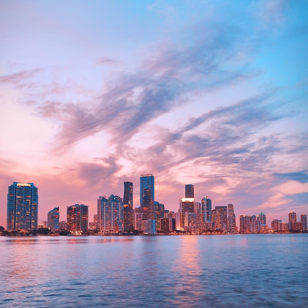 An epic cityscape shot of Miami at dusk taken from the water. The sky is filled with cotton-candy fluffy cloud wisps in soft shades of cool pink and soft blue.