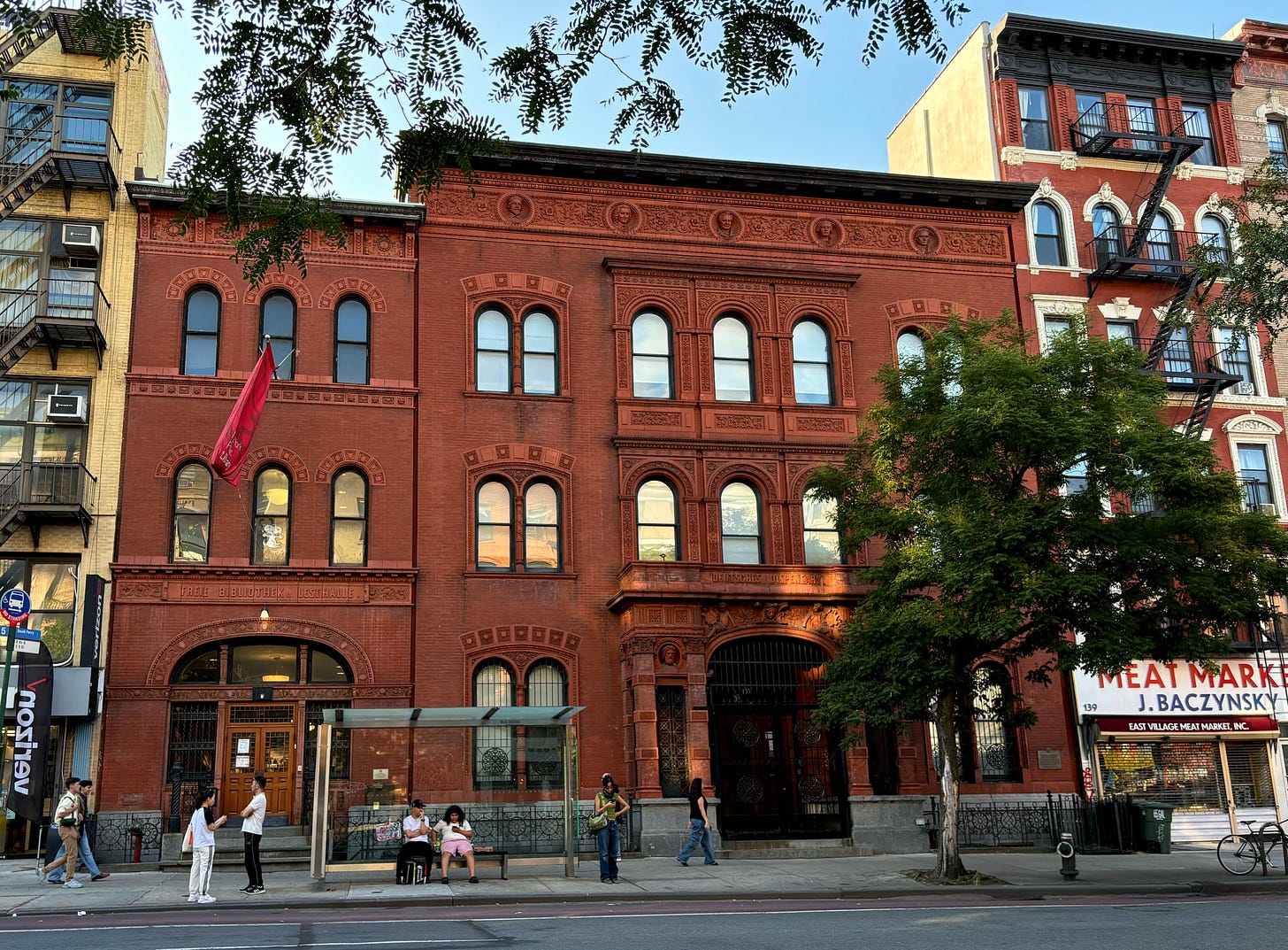 Two ornately decorated red terra-cotta buildings, the one of the right slightly taller and wider than its twin on the left. The one on the left has a red NYPL flag outside. The one on the right has a decorated portico.