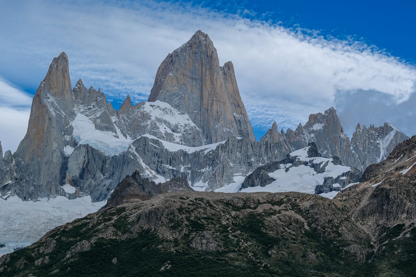 The Fitz massif, comprised of several granite spires and peaks, rise from the Patagonian forest floor, still covered in snow. Fitz Roy is the most prominent in the center, rising against a cloud-filled sky.