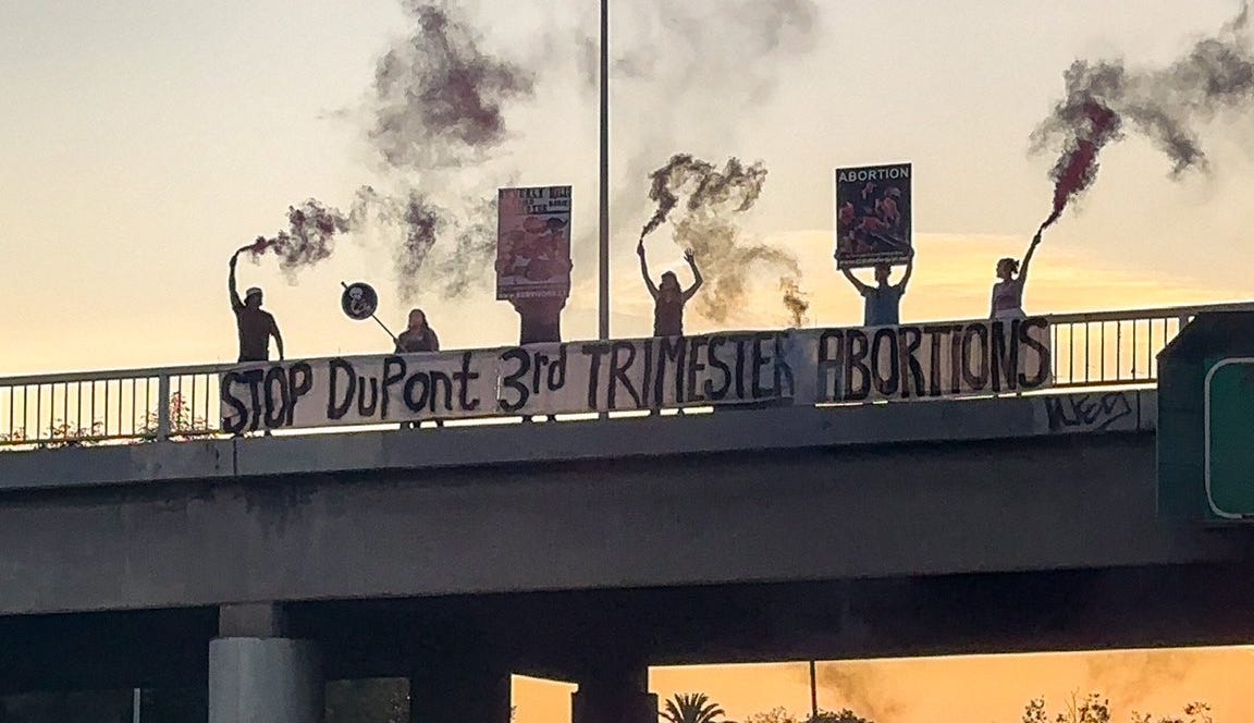 People on a bridge over a highway holding a banner that reads Stop DuPont 3rd Trimester Abortions