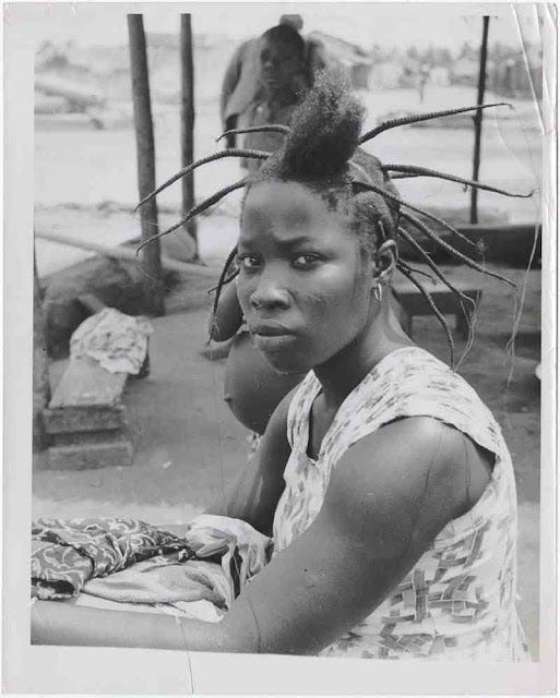 black and white photograph taken by Richard Wright on his trip to Ghana pre-independence (circa 1953). A Ghanian woman has her hair threaded except for a small portion at the top of her head, near her forehead. She wears a simple cotton dress with a repeating pattern and looks directly into the camera over her shoulder. Two people stand behind her in the background: a child who looks over at the cameraman and off, to something unseen. A round and protrudent belly juts out from the behind the woman with threaded hair, with their face obscured.