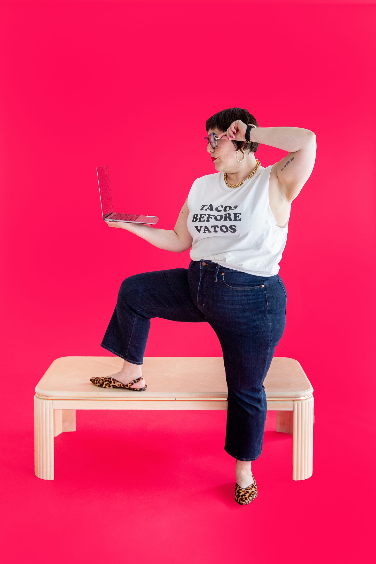 Lauren stands with one leg up on a table looking over at her computer that's in her left hadn while her right hand is on her glasses. Her shirt reads "tacos before vatos"