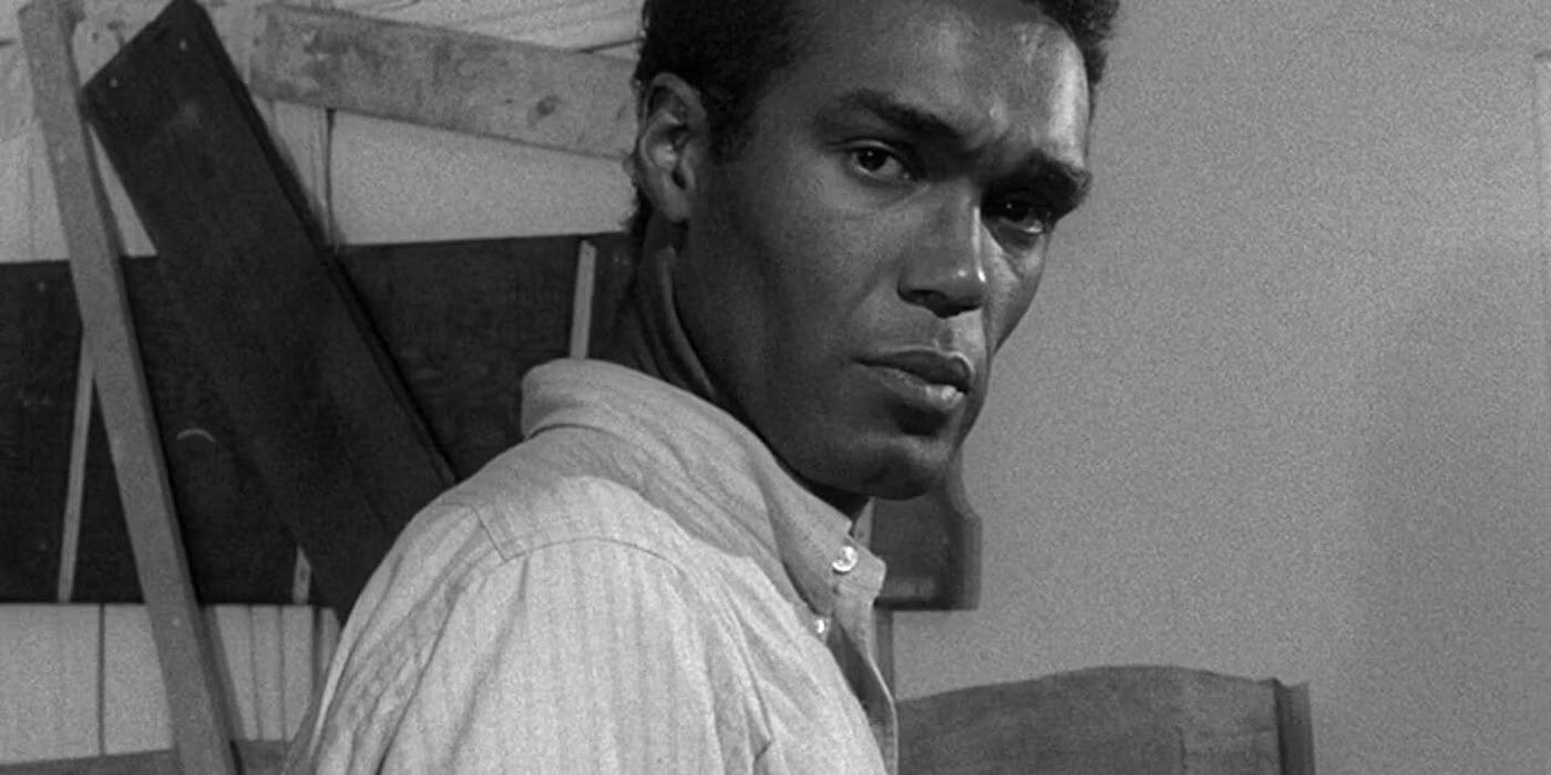 Black and white screenshot of Night of the Living Dead. A handsome Black man (Duane Jones as Ben) in a shirt looks at the camera with a serious expression. Behind him, a boarded window.